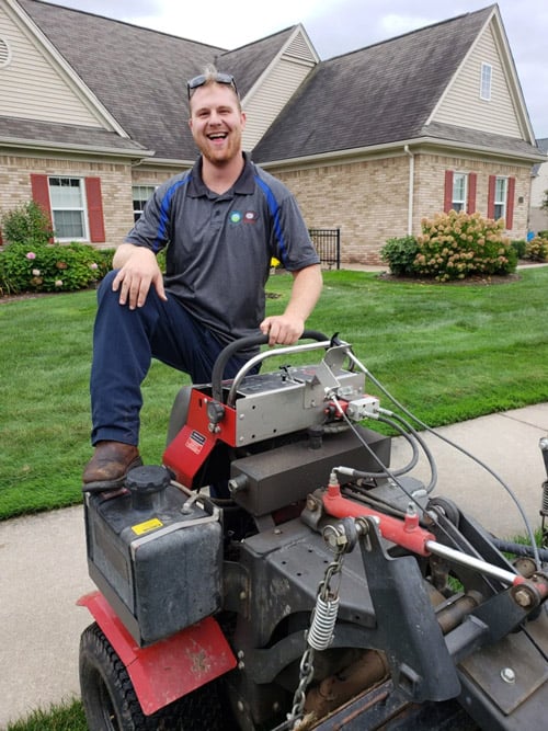 Lush Lawn franchise technician smiling next to professional lawn care equipment in a residential neighborhood